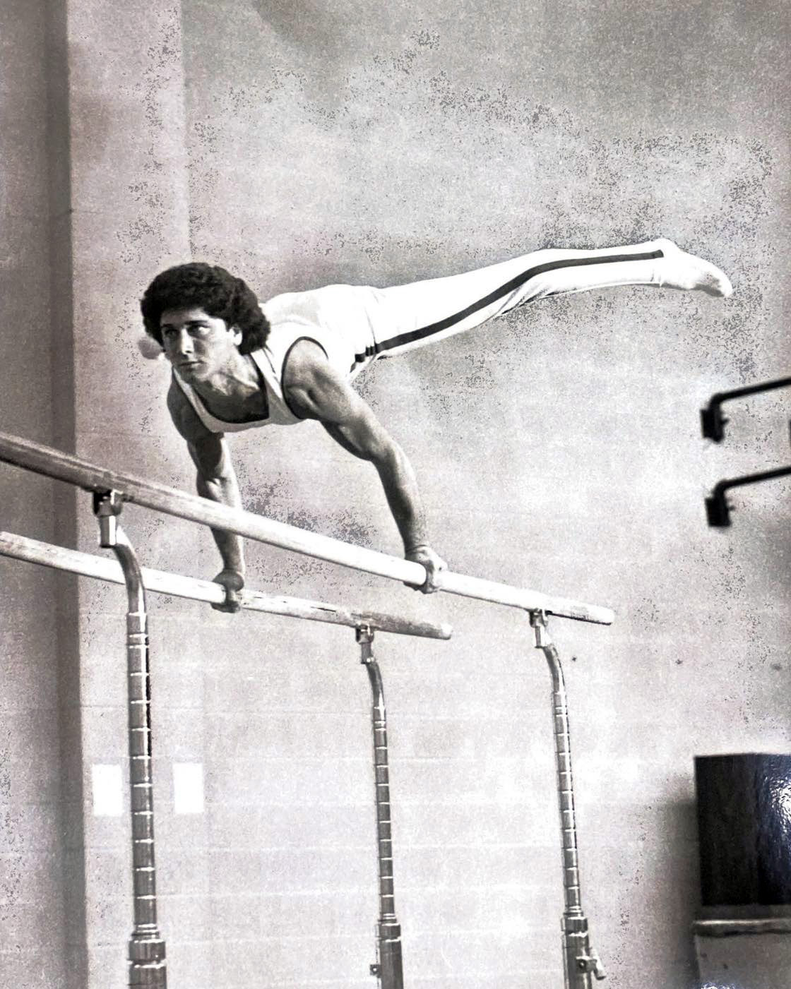 A black and white photos of a man holding a planche balancing position on the parallel bars. 