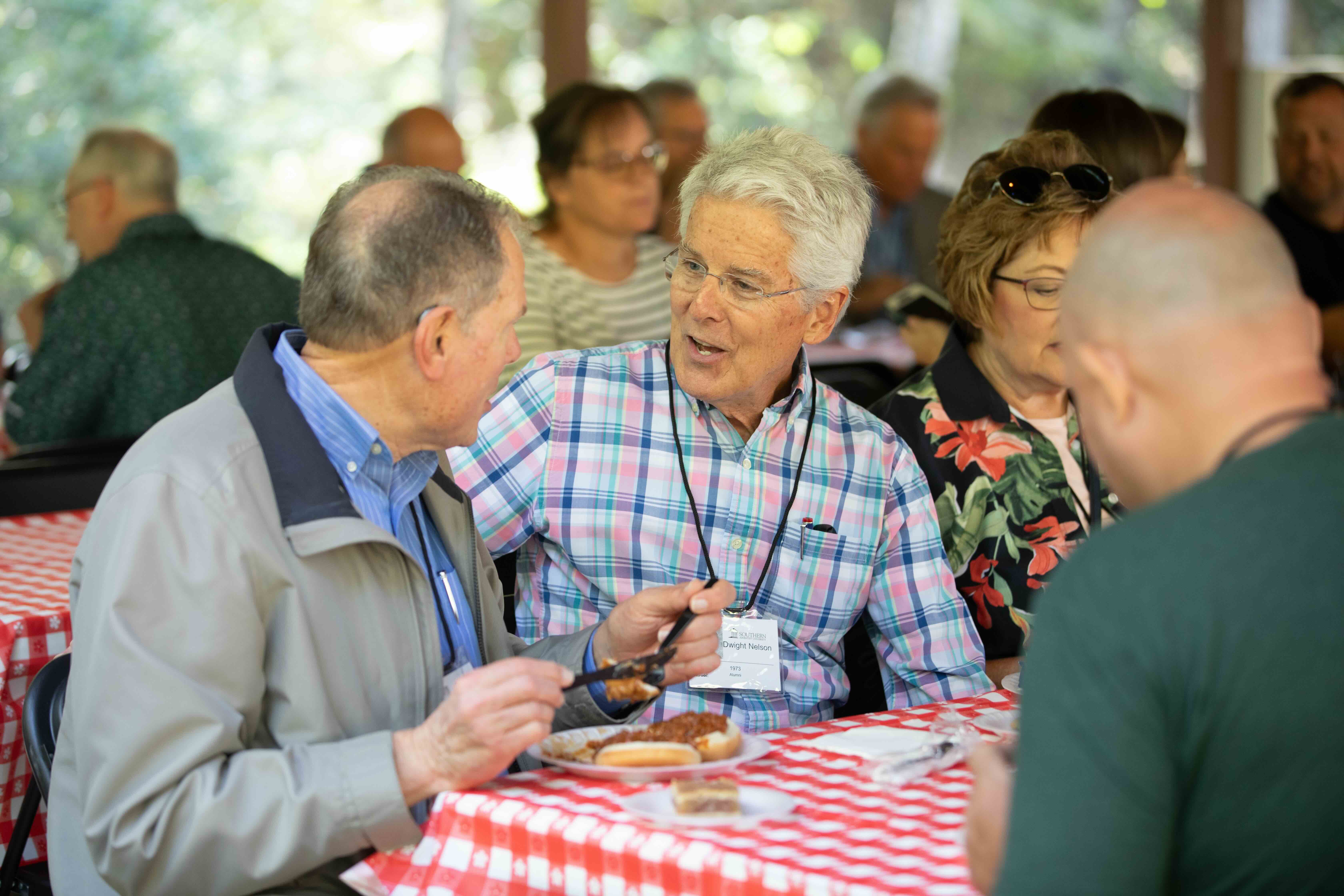 Homecoming Weekend Photo of 2 alumni eating lunch together at a picnic table