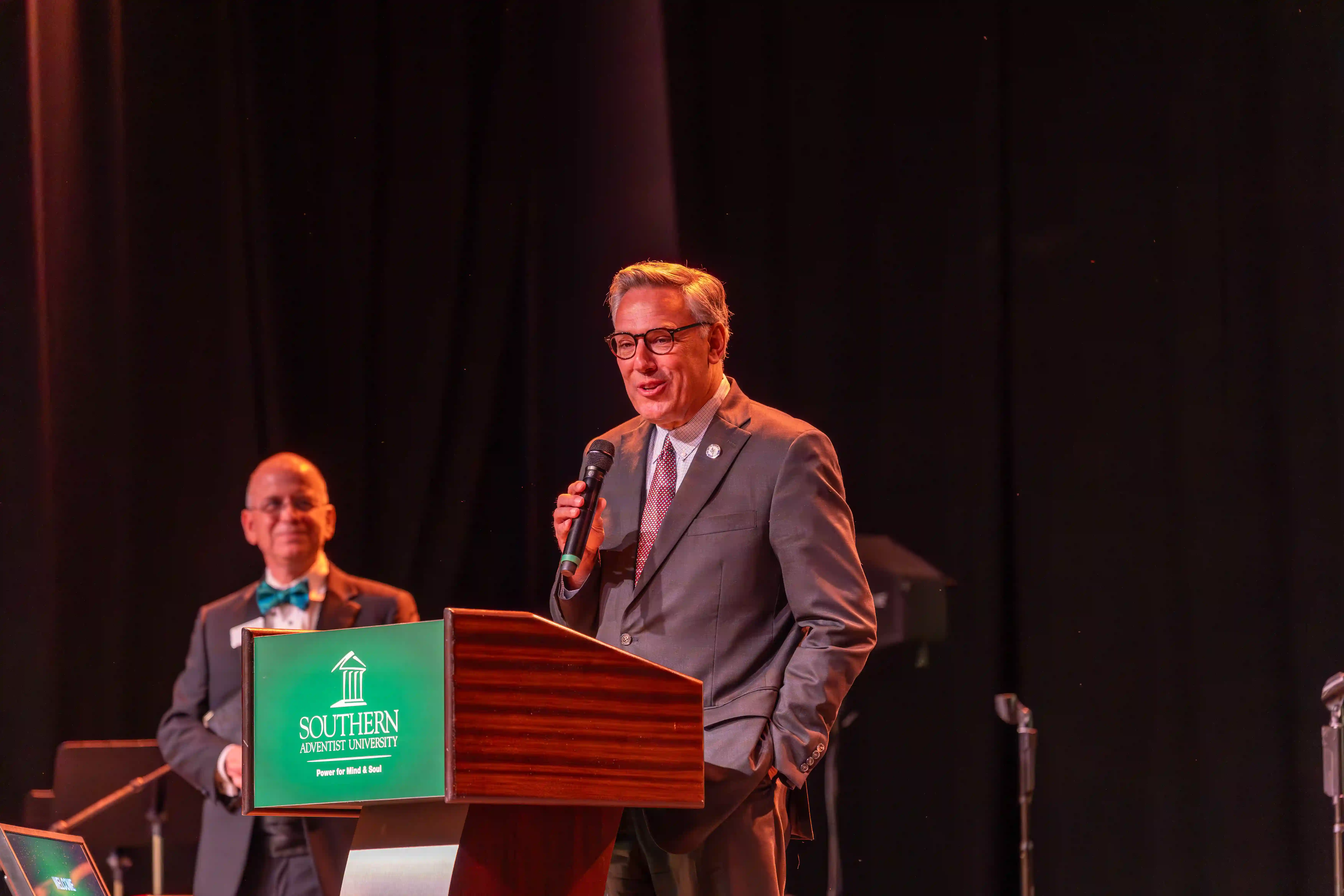 Greg Vital speaking at the Southern Adventist University Podium while Ken Shaw watches from farther back on the stage